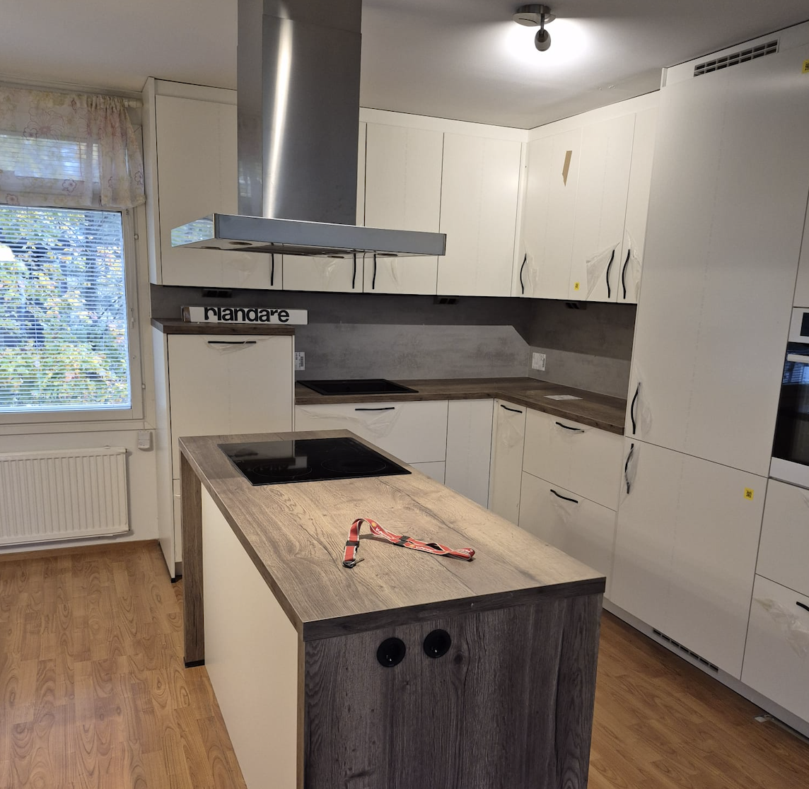 Moderni keittiö valkoisilla kaapeilla, harmaalla tasolla ja liesitasolla, ikkunan edessä puulattia.